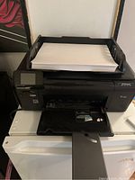 Black HP Photosmart all-in-one printer loaded with white paper in rear feed tray, front output tray extended, touchscreen and cartridge access visible