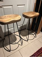 Pair of solid wood stools with contoured seats and black metal legs placed on tiled floor.