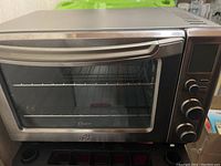 Front view of Oster toaster oven showing glass door, wire rack inside, and control knobs with digital display.