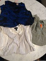 Three women's silk tops displayed on a rounded marble table: blue and black sleeveless top, cream sleeveless top with stripes, and greenish-gray patterned long sleeve top.