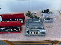 Wide view showing two Proto red metal socket cases, a green hand planer, a gray plastic socket set case, small plastic containers with parts, and wrench extensions spread on a beige surface.