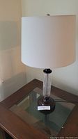 Front view of the table lamp showing the white drum lampshade and pinwheel crystal stem mounted on a dark base, placed on a wooden and glass side table.