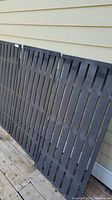 Three dark-colored privacy panels with vertical slats leaning against a beige exterior wall on a wooden deck.
