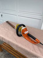 Full side view of the Stihl HSE 70 electric hedge trimmer showing orange and black handle with attached cord and long dual-sided blade lying on a pink cloth surface.