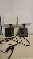 Two black wall lamps side by side on table, showing bases, arms, and white square shades with cords tangled in front.