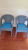 Front view of the two gray wicker chairs with blue cushions placed side by side on a wood floor against a white wall.