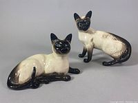 Two Beswick porcelain cat figurines, one standing and one lying, both painted in Siamese coloring, placed on a grey background.