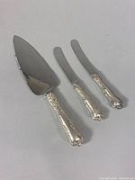 Full view of the cake cutter and two butter spreaders showing sterling silver handles with ornate floral pattern and stainless steel blades.