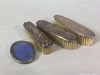 Four clothing brushes, three sterling silver with various designs and monograms, and one glass frame with blue backing and silver edge with patina and dents visible.
