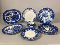 Assorted blue and white Johnson Bros Normandy Flow Blue china pieces including oval serving platters, round plates with floral and scenic patterns, and a teacup with saucer.