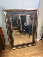 Front view of the rectangular framed beveled mirror leaning against a wall on a wooden floor showing the full decorative frame.