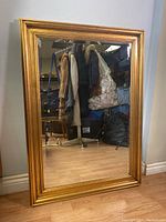 Full view of rectangular beveled mirror with gold frame against a wall, reflecting some coats and bags.