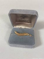 Gold leaf-shaped pin inside grey velvet Maurice Ltd. box, showing the front with intricate leaf design and fine detailing.