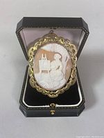Front view of gold framed cameo brooch in black box, showing detailed carving of woman painting on easel.