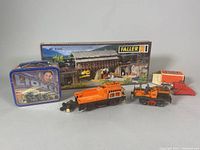 Photo shows entire lot including Lionel tin lunch box, Faller B-535 boxed bridge, orange train bumper, small orange vehicle with figure, and small additional train parts.