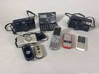 Front view of all items including three film cameras, five mobile phones, a remote control, and a rotary phone keypad accessory on a white surface.
