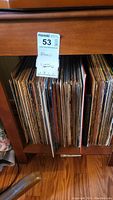 Photo of wooden shelf holding numerous vintage vinyl albums tightly packed, label tag with lot number visible.