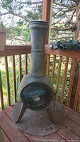 Full front view of tall cast metal chiminea on fireproof deck base, standing on an outdoor wooden deck with trees visible in background.