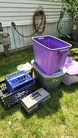 Full view of the items including purple and grey plastic totes, black milk crates, grey cleaning bucket, and toolbox on grass outside near house wall.