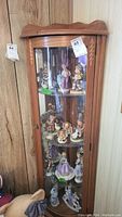 Front view of wooden corner curio cabinet filled with various figurines (contents not included). Shows carved details and curved top molding.