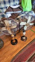 Two vintage electric fans placed on a wooden floor in front of a couch. One taller fan on the left and one smaller fan on the right. Both have metal blades and wire cages.
