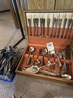 Photo showing one open box with metal flatware stored in orange case, including forks, spoons, serving spoons with decorative cutouts, and ladles; next to it is a plastic container with wooden handled utensils.