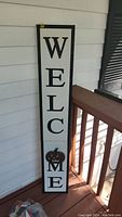 Tall wooden Welcome sign painted white with black letters and a decorative sunflower near the bottom leaning against a white wall with wood siding and a porch railing