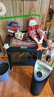 Black electric heater stove with glass front panel and a decorative clock and doll on top. Floor and wooden panel backdrop visible.
