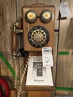 Wooden TeleConcepts rotary dial phone with dual metal bells and handset attached. Phone is mounted on a wooden board with a ledge and a coiled white cord. Instruction manual is placed on ledge below phone.