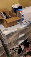 Photo of 3 drawer plastic storage unit with items on top and inside drawers including cardboard boxes and various cords.
