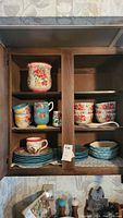 Cabinet with colorful floral mugs, small floral bowls, turquoise dinner plates, and a large floral patterned cup or jar on top shelf.
