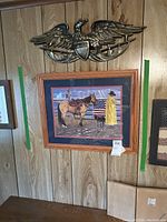 View of framed cowboy artwork and metal eagle crest wall hanging on wood-paneled wall