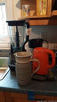 Ninja blender with clear pitcher on base, orange electric kettle, and pottery jug on kitchen counter near window and sink.