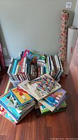 View of a large assortment of children's books and cookbooks stacked on a hardwood floor near a wall.