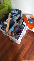 Plastic bins stacked containing various crafting materials including a wooden T-square ruler and purple yarn ball
