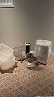All items in lot including plastic storage bins, metal mesh waste basket, small desk fan, Lasko oscillating fan and Whirlpool Gold dehumidifier displayed on carpeted floor