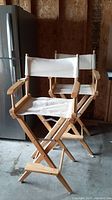 Two bar height wooden director chairs with white canvas seats and backs shown side by side in garage with concrete floor and unfinished wall. One chair is highlighted in the foreground with armrests and foldable wood frame.