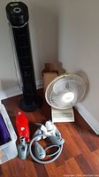 Wide shot showing Ceville black tower fan, Windstream white table fan with control buttons, Dirt Devil vacuum body, Shark vacuum hose and accessories on wooden floor.
