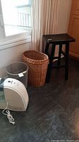 Wide shot showing the Honeywell humidifier on the floor next to a large wicker basket with a mesh basket insert and a black wooden stool near the corner of the room.
