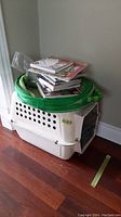 White plastic pet carry cage with black metal door and green collapsible mesh tube carrier on top, and various books stacked on top of the carriers.