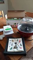 Photo showing Betty Crocker popcorn maker, RTMAXCO veggie gadget box and device, metal napkin holder with weighted arm and napkins on wooden table.