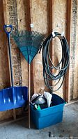Shovel, rake, garden hose coiled on holder, blue bin with bags inside in garage against wood panel wall