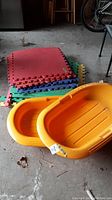 Photo showing two orange plastic sleds next to a stack of colorful foam interlocking floor mats.