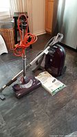 Kenmore canister vacuum cleaner with burgundy body, upright handle attachment, metal wand, orange power cord wrapped around handle, and vacuum bags on the floor.