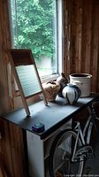 Washboard standing next to three pottery jugs and one white crockery pot on table near window