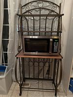 Front view of metal bakers rack with wood shelves and microwave on middle shelf, decorative scrollwork.