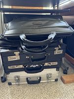 Four briefcases stacked: top is black leather Bugatti satchel, below three hard-sided briefcases in black and silver with combination locks and handles.