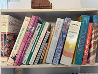 Cookbooks arranged upright on a white shelf with spines facing forward showing various titles focused on cooking techniques and recipes.
