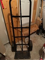 Front view of the black metal moving dolly showing the vertical support bars, flat loading plate, and one tire.