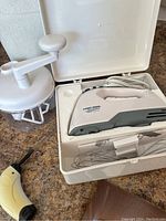 Image of manual hand operated mixer with clear plastic mixing container and white handle next to closed case with electric mixer and immersion blender.